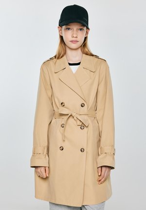Młoda osoba w beżowym płaszczu dwurzędowym z paskiem wiązanym w talii i czarnej czapce z daszkiem, stojąca na tle jednolitego jasnego tła.