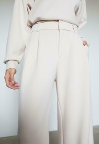 Beige brede bukser lavet af glat stof, med høj talje, folder og sidelommer. Benene er løse og svævende.