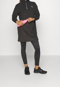 Zwarte fleece jurk met halve ritssluiting en zijzakken, gecombineerd met zwarte-gepatroneerde leggings en zwarte sneakers, terwijl ze een roze waterfles vasthoudt.