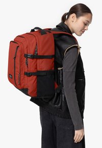Mochila roja con detalles en negro, hecha de un tejido duradero, que cuenta con correas ajustables, múltiples bolsillos y tiradores de cremallera en contraste.