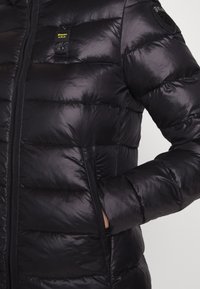 Sort pufferjakke med blank finish, fremhævet af vandret quiltet mønster, sidelommer og logo patch på venstre bryst.
