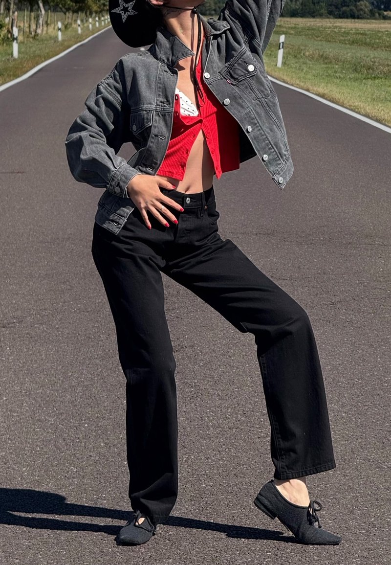 Veste en denim gris portée sur un top rouge court, pantalon noir à jambes larges, et chaussures noires à nouer sur le devant, posée devant un paysage routier.