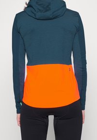 Sportig jacka med en turkos övre sektion och en ljus orange nedre sektion. Huva med dragkedja på sidan.