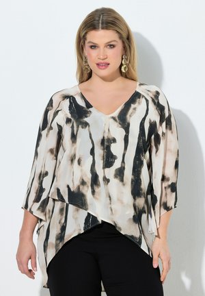 Kobieta o blond włosach, nosząca złote kolczyki oraz czarno-białą, luźną bluzkę w wzory z rękawami 3/4 i czarne spodnie na białym tle.