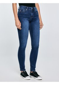 Sieviete tērpusies tumši zilās pieguļošās džinsu biksēs un melnās zema profila sporta kurpēs, stāv pret vienveidīgu gaiši pelēku fonu.