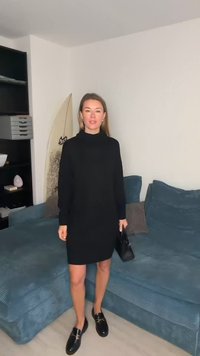 Juoda, ilgomis rankovėmis megzta suknelė ilgumu iki kelių su aukštu kaklu, derinama su blizgiais juodais bateliais be kulno ir maža rankine, fone – mėlynas sofa.