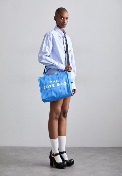 Sac fourre-tout bleu avec texte blanc, doté de poignées plates et de forme rectangulaire. Le modèle porte une chemise bleu clair et des chaussures noires avec une sangle.