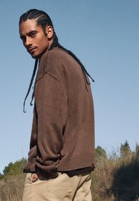 Brun stickad tröja med ribbat fåll, buren över en svart skjorta. Modellen har flätat hår och står utomhus mot en klar himmel.