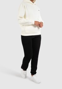 Cremefarbener Hoodie mit einer Fronttasche und einem Kordelzug, kombiniert mit schwarzen Jogginghosen. Weiße, strukturierte Sneaker vervollständigen das Outfit.