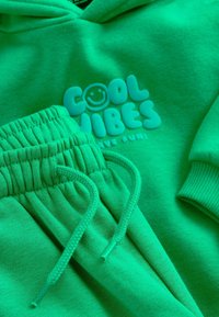 Zöld kapucnis pulóver és melegítő szett. Puha anyagból, "COOL VIBES" felirattal és mosolygós arccal díszítve. Bordázott mandzsetták és állítható derékú, húzózsinóros szegéllyel.