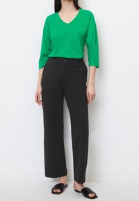 Grünes V-Ausschnitt Langarmtop kombiniert mit hochtaillierten schwarzen Hosen. Glatter Stoff, lockere Passform und schwarze offene Mules rundet das Outfit ab.