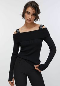 Črn pulover z dolgimi rokavi z rebrastim vzorcem in odkritimi rameni, pridobljenimi naramnicami z zakovicami, oprijet kroj, kombiniran s črnimi hlačami.