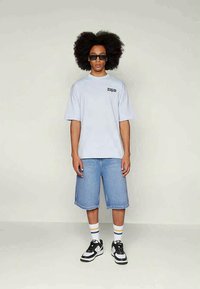 T-shirt oversized azul claro com logótipo, calções de ganga azuis, sapatilhas pretas e brancas, e meias riscas. Modelo em pé com as mãos à mostra.
