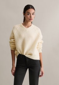 Jasnokremowy dzianinowy sweter z dekoltem w serek, fakturowanym wzorem i luźnym krojem, zestawiony z czarnymi dżinsami.