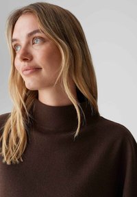 Bruine gebreide trui met hoge kraag, met een zachte textuur. Het model heeft golvend, schouderlang haar en een neutrale achtergrond.