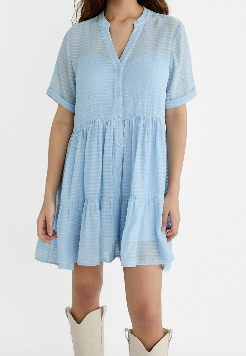 Femme portant une robe courte boutonnée bleu clair texturée avec des manches courtes et des bottes de cowboy crème.