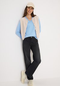 Lichtblauwe v-hals trui, gelaagd onder een crèmekleurig, knoopjesvest. Zwarte loszittende broeken en beige sneakers maken de outfit compleet.