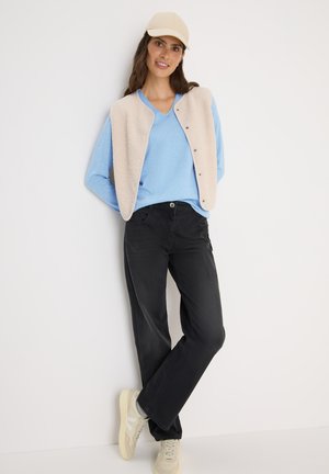 Un pull bleu clair en V superposé sous un gilet crème à boutons. Un pantalon noir ample et des sneakers beiges complètent la tenue.
