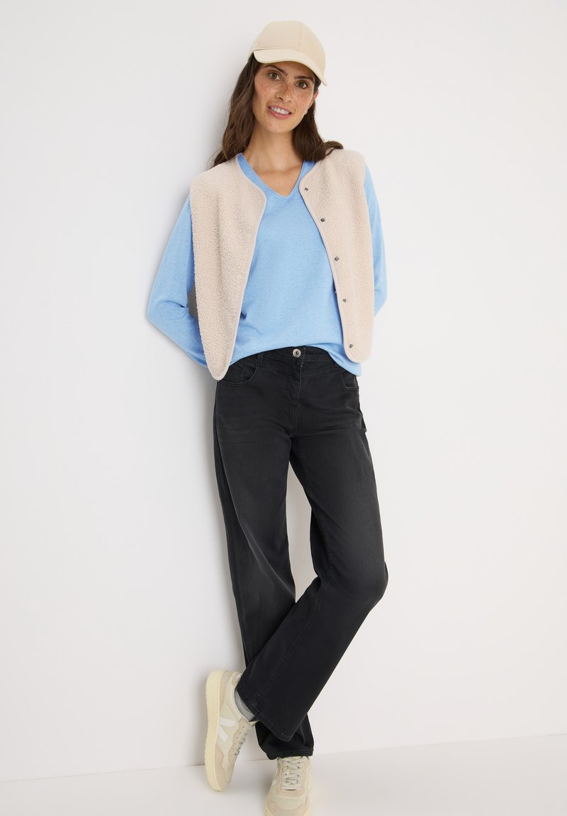 Lichtblauwe v-hals trui, gelaagd onder een crèmekleurig, knoopjesvest. Zwarte loszittende broeken en beige sneakers maken de outfit compleet.