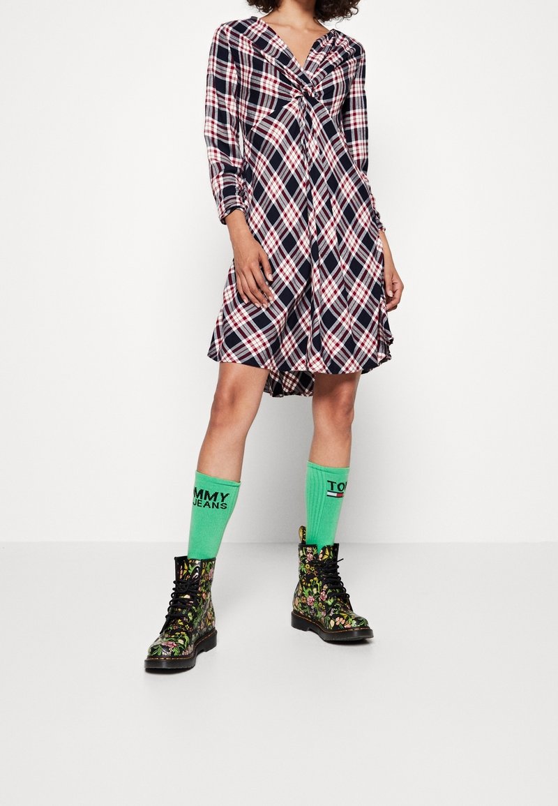 Femme portant une robe à carreaux noire, blanche et rouge longueur genou, avec des chaussettes vertes Tommy Jeans et des bottines noires à lacets à motif floral.