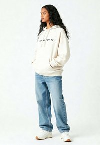Felpa crema con cordini e tasca frontale, con testo nero. Abbinata a jeans azzurri a vestibilità ampia e sneakers bianche.
