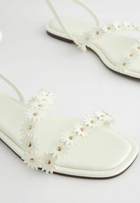 Weiße flache Sandalen mit quadratischem Zehendesign, verziert mit floralem Applikationen und goldenen Nieten entlang der Riemen. Glatte Textur und minimalistischer Stil.