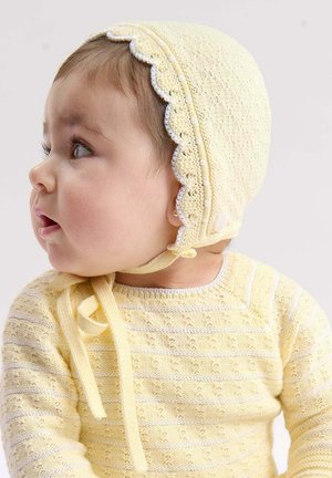 Baby in een gele gebreide muts met geschulpte randen en bijpassende gele gebreide trui, kijkend naar de zijkant tegen een neutrale achtergrond.