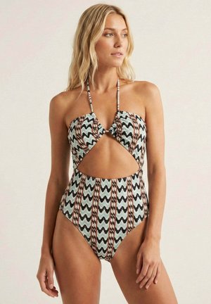 Mujer rubia con un bañador de una pieza estampado con cuello halter y abertura en la cintura, mirando hacia un lado sobre un fondo neutro.