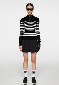 Svart och vit randig långärmad tröja med dragkedja vid kragen, kombinerad med en svart plisserad kjol. Svarta sneakers och knähöga strumpor med detaljer.