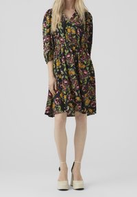 Robe fleurie en tissu noir avec des motifs roses et verts, présentant une taille froncée, un décolleté en V et des manches trois-quarts. Portée avec des chaussures à plateforme beiges.