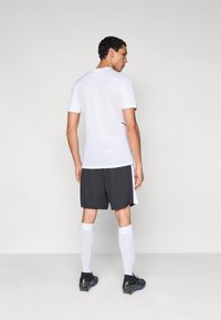 Fehér sportpóló rövid ujjakkal, fekete díszítéssel; sötét rövidnadrággal, fehér zoknival és fekete focicipővel párosítva. Hátsó nézet.