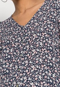 Blusa floreale blu navy con scollatura a V, maniche corte e piccoli bottoni, con un motivo di fiori rosa e bianchi su uno sfondo scuro.