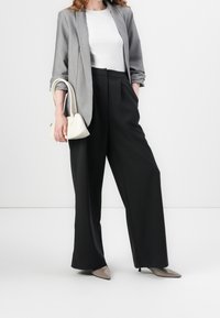 Šedý blazer přes bílou košili, černé kalhoty s širokými nohavicemi a béžové kožené lodičky na špičce. Malá bílá kabelka dotváří celý outfit.