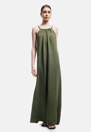 Donna che indossa un vestito lungo senza maniche di colore verde oliva stropicciato con fiocchi sulle spalle e sandali neri su uno sfondo chiaro uniforme.