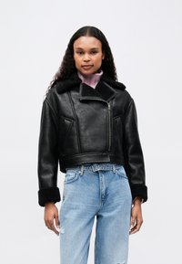 Γυναίκα με σγουρά μαλλιά που φοράει μια μαύρη γούνα πάνω από ένα ροζ ζιβάγκο και ανοιχτό μπλε σκισμένο τζιν σε απλή φόντο.