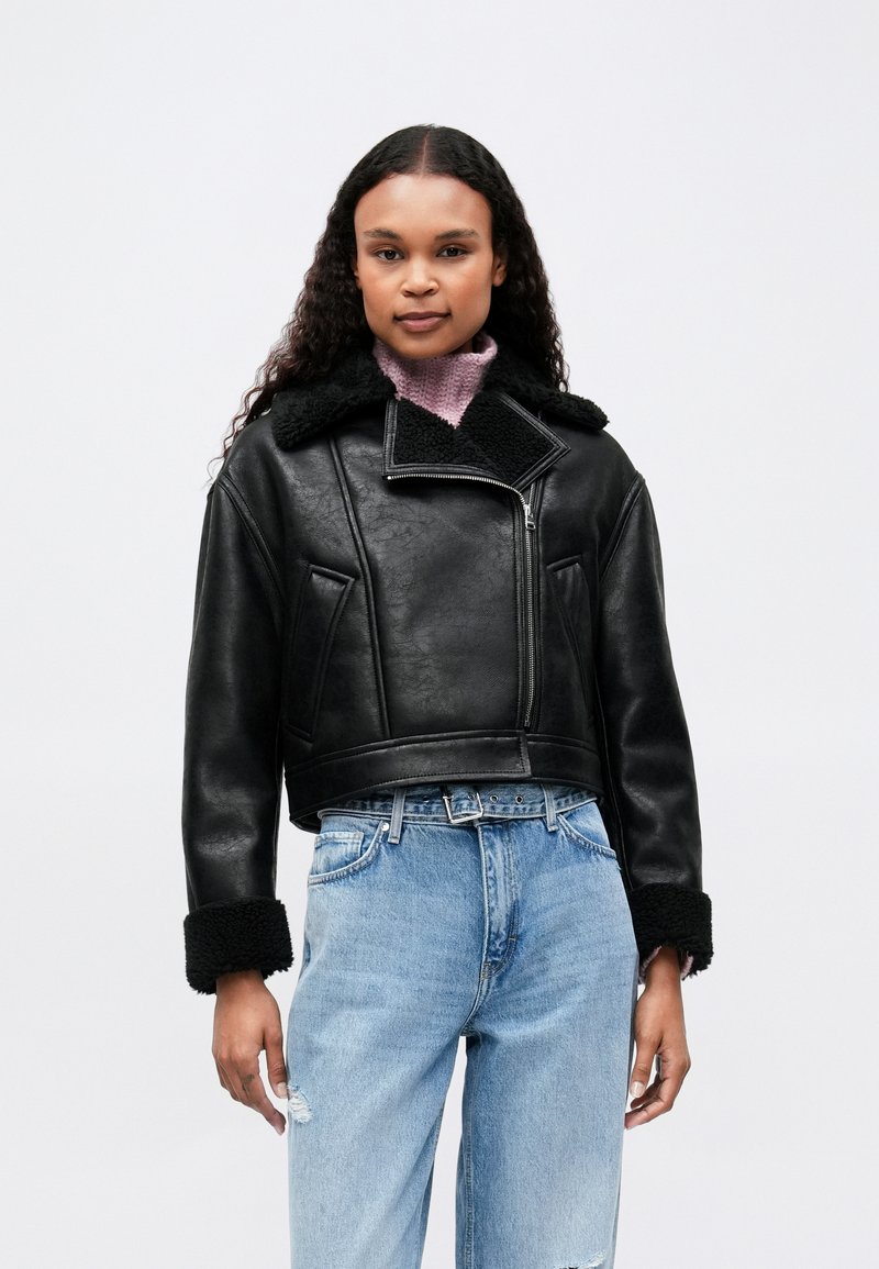 Γυναίκα με σγουρά μαλλιά που φοράει μια μαύρη γούνα πάνω από ένα ροζ ζιβάγκο και ανοιχτό μπλε σκισμένο τζιν σε απλή φόντο.