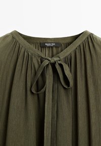 Blouse froncée vert olive avec un col rond et un nœud noué à l'avant, avec une étiquette Massimo Dutti visible à l'intérieur du col.