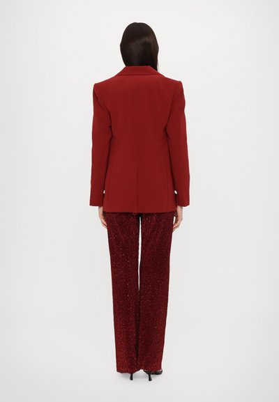 Rdeč blejzer z oblikovanimi rameni in odprtim ovratnikom, v kombinaciji z bleščečimi rdečimi širokimi hlačami. Gladek material, elegantna zasnova in sijoč detajl.