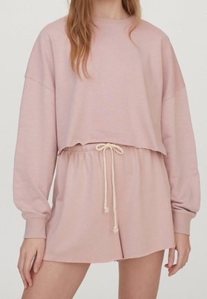 Femme portant un sweat-shirt court à manches longues rose clair et un short taille haute assorti avec un cordon blanc.