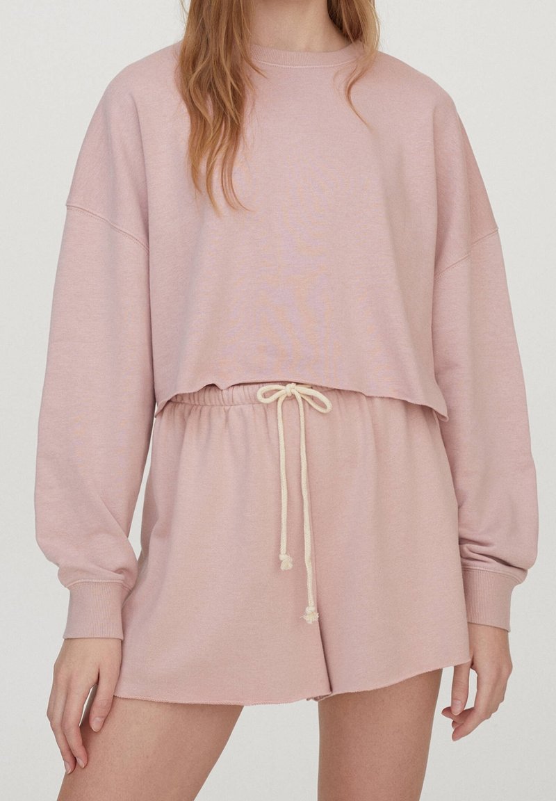 Femme portant un sweat-shirt court à manches longues rose clair et un short taille haute assorti avec un cordon blanc.
