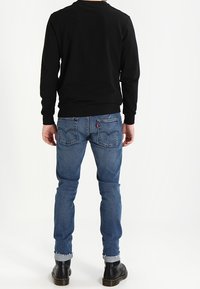 Sudadera negra de manga larga y jeans azules desgastados con corte recto, dobladillo enrollado y botas negras con cordones. Detalle visible de costura en el bolsillo trasero.