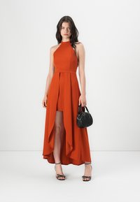 Rust-orange halter neck dress features a high-low hem, pleated detailing, and a flowing design; paired with a small black handbag and heels.