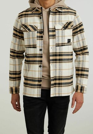 Man wearing beige hoodie under a beige, black, and white plaid button-up shirt with black pants.