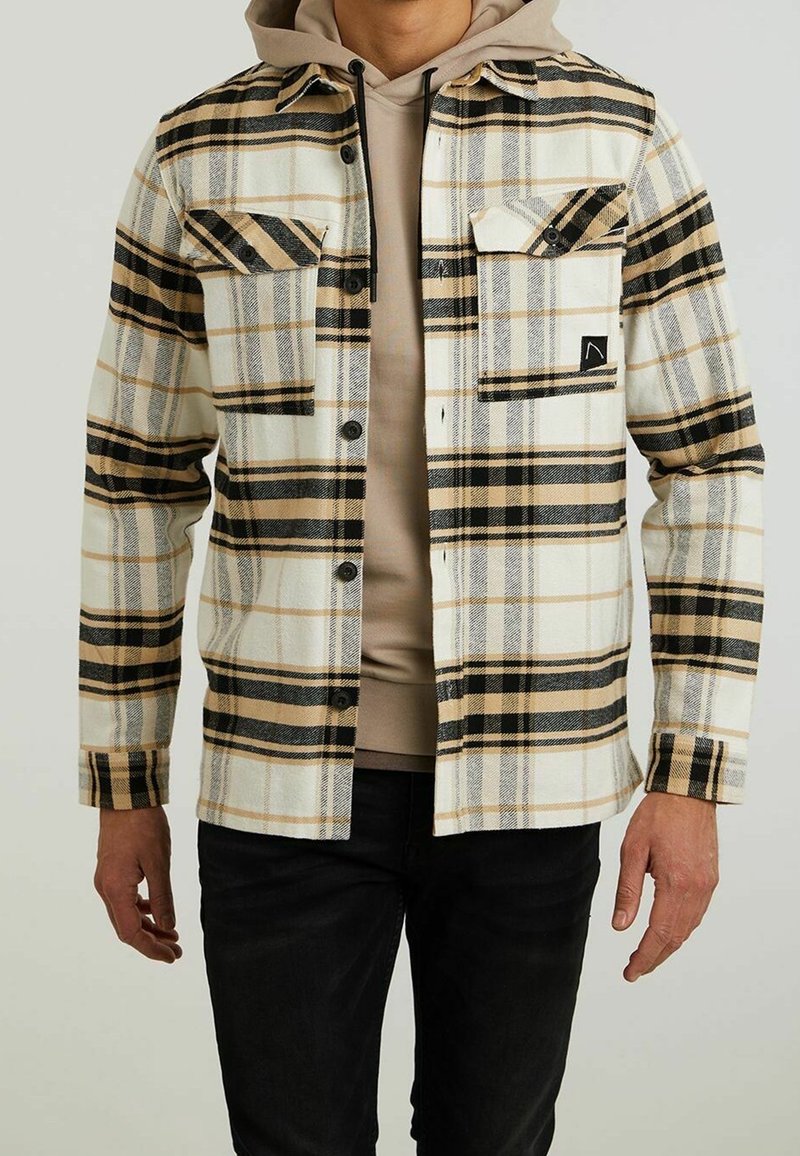 Homme portant un sweat à capuche beige sous une chemise à carreaux beige, noire et blanche boutonnée avec un pantalon noir.