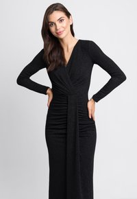Czarna, długa sukienka z długim rękawem i krojem kopertowym, z marszczeniami w talii, wykonana z błyszczącego materiału, sięgająca do ziemi o gładkiej fakturze.