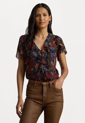 Blusa floreale in blu scuro con dettagli arricciati, decorata con fiori rossi, blu e gialli. Abbinata a pantaloni marroni a vita alta e una cintura.