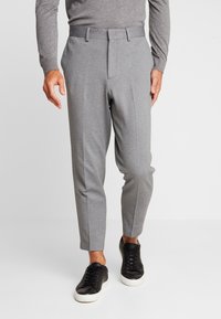 Grå byxor med avsmalnande passform, med en platt frontdesign och subtil textur, kombinerade med svarta sneakers. Detalj med elastisk midjeresår är synlig.