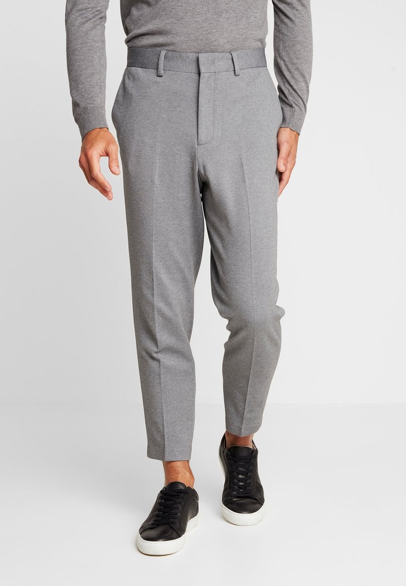 Grå byxor med avsmalnande passform, med en platt frontdesign och subtil textur, kombinerade med svarta sneakers. Detalj med elastisk midjeresår är synlig.