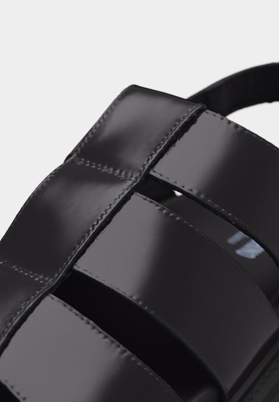 Sandale en cuir noir avec des larges brides lisses et des surpiqûres visibles, montrant un gros plan du détail du tissage.