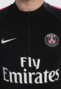 Schwarzes Sporttrikot mit halbem Reißverschluss. Mit pinken Akzenten an den Schultern und weißen Logos: "Paris Saint-Germain" und "Fly Emirates." Hergestellt aus atmungsaktivem Material.
